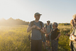 Eine Person mit einem Basecap steht lächelnd in der Mitte des Bildes auf einer saftig grünen Wiese mit hohem Gras. Am Rand und im Hintergrund sieht man weitere Personen die in die Gegend blicken.