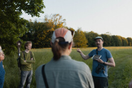 n der Bildmitte ist unscharf eine Person mit Zopf und Cappy von hinten zu sehen. Weiter im Hintergrund sieht man zwei Menschen links und rechts von ihr. Auf der linken Seite steht ein Mann mit verschränkten Armen und hört dem anderen Mann auf der rechten Seite zu, der gerade etwas auf einem Tablet zeigt und dazu mit dem Finger gestikuliert. Der Hintergrund ist wieder von einer satt grünen Wiese und einem Laubbaumrand bestimmt.